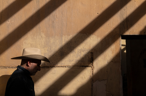 Side view of adult man in cowboy hat against wall with sunlight and shadow