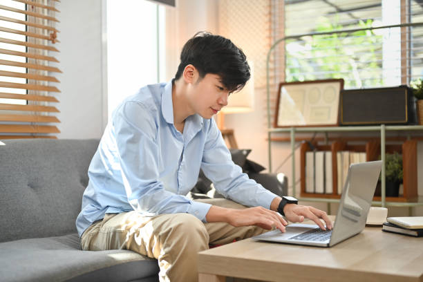 istock Happy young asian man browsing internet, booking hotel online on laptop while sitting on sofa at home. 1467807458