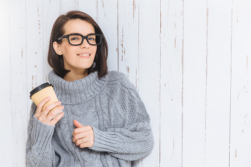 Pretty brunette young female in knitted warm sweater, holds hot drink in paper cup, tries to warm herself in cold winter day, isolated over white background. Adorable woman has coffee break.