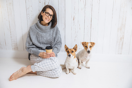 Indoor shot of beautiful woman wears spectacles and casual wear, drinks hot beverage, enjoys good morning, sits near her favourite pets against white fence background. Happiness and leisure concept