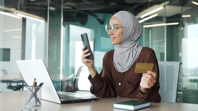 Positive young muslim businesswoman in hijab paying with credit card on smartphone at office workplace
