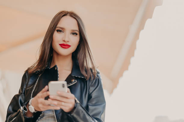 Horizontal shot of attractive dark haired female with red painted lips, dressed in black leather jacket, holds modern cell phone, sends text messages, chats online, strolls at city street. Technology tiro horizontal de atractiva mujer de pelo oscuro con labios pintados en rojo, vestido con chaqueta de cuero negro, sostiene el teléfono celular moderno, envía mensajes de texto, chats en línea, paseos en la calle de la ciudad. tecnología
