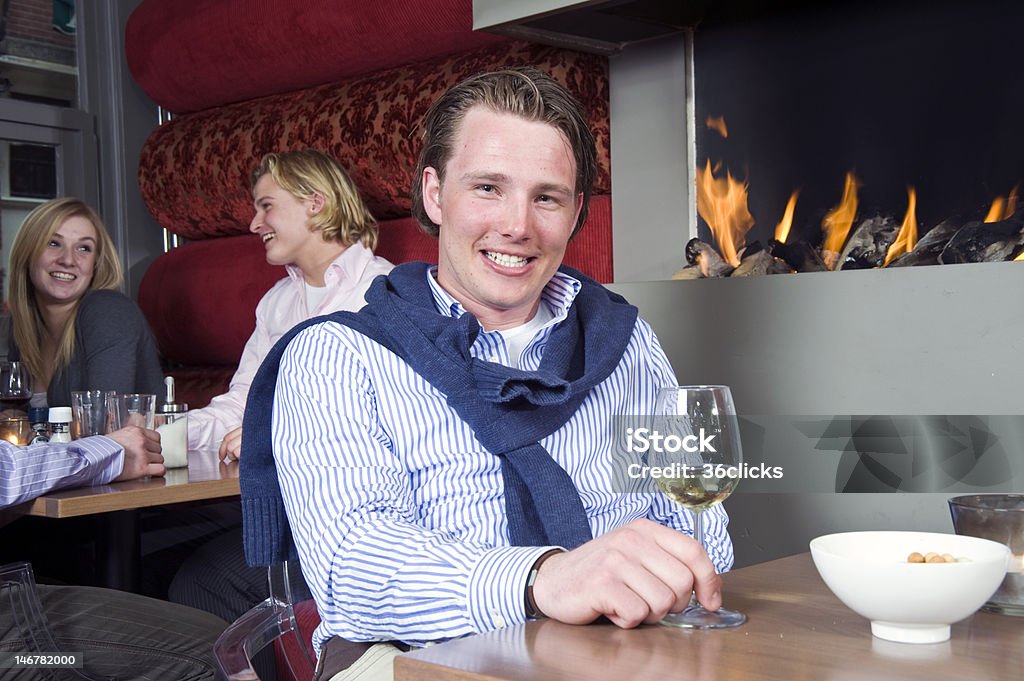 Posh Dandy A posh looking man with his sweater around his neck and a glass of white wine in his hand sitting at a restaurant table in front of the fireplace Yuppie Stock Photo