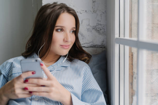 la donna bruna premurosa ascolta l'audiolibri, tiene in mano lo smartphone moderno, gode di un suono perfetto negli auricolari guarda pensierosamente alla finestra vestita con abiti casual. la femmina ascolta la musica nella playlist - domestic staff audio foto e immagini stock