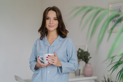 Morning time concept. Young pretty European woman in nightclothes carries white mug of coffee or cappuccino, stands indoor in living room, has weekend, enjoys good rest. People and lifestyle