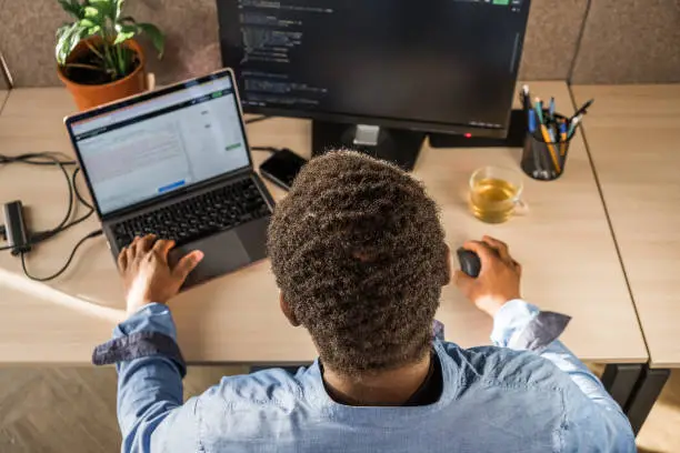 Black Man Writing Lines of Code On Desktop PC With Two Monitors and a Laptop Aside in Stylish Office. Professional Male Developer Programming Artificial Intelligence Software for Start-Up Company.