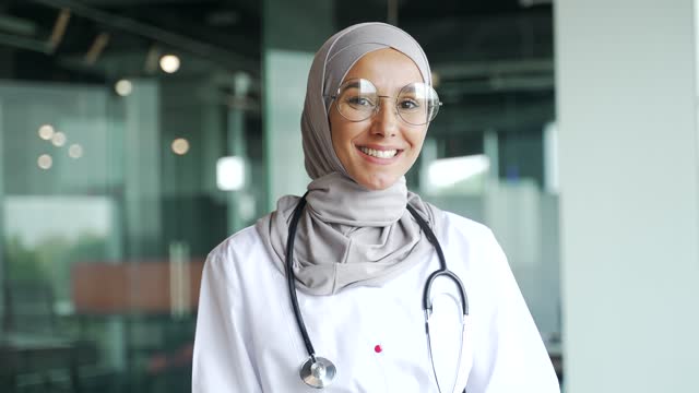Portrait of smiling young muslim doctor therapist in hijab with stethoscope looking at the camera in glass workplace Happy medical worker