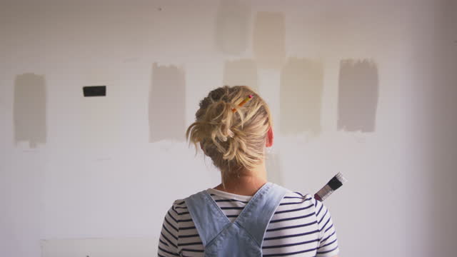 Rear View Of Woman Decorating Room In House Painting Tester Paint Colour Strips On Wall