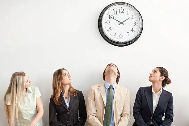 Four businesspeople sitting on bench and looking up at clock. Horizontally framed shot.