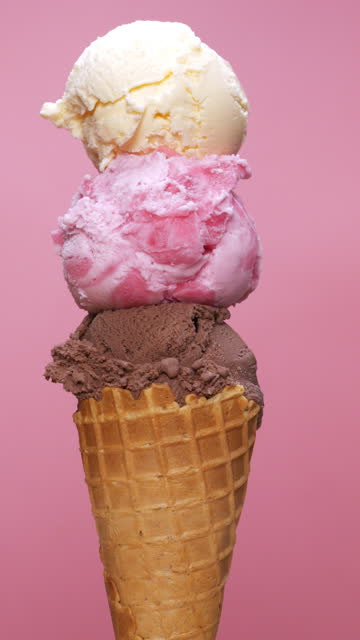 Vertical video front view, Scooping vanilla ice cream on top of strawberries and on a chocolate ice cream cone on a pink background.