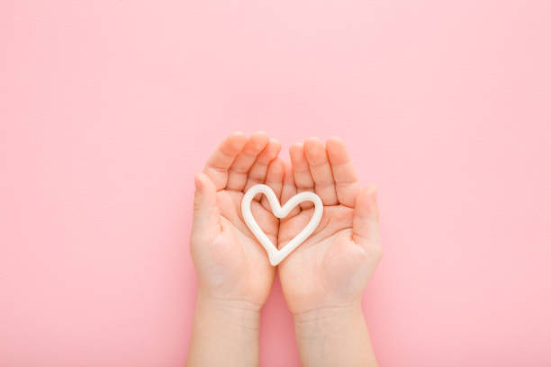White heart shape on little baby girl opened palms on light pink table background. Pastel color. Closeup. Point of view shot. Love concept. Care about child hands skin. Top down view. forma de corazón blanco en las palmas de las palmas de la niña abiertas sobre el fondo de la mesa de color rosa claro. color pastel. closeup. toma de punto de vista. concepto de amor. cuidado de la piel de las manos del niño. vista de arriba hacia abajo