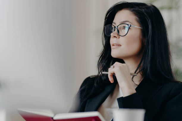 istock Thoughtful European woman makes notes in notepad, holds pen and writes down some ideas, makes list of goals, plans day in personal organizer, wears transparent glasses, thinks about something 1467849637