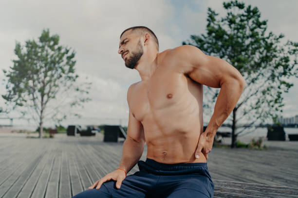 displeased unshaven european sportsman touches lower back and feels backache after physical training exercises, poses outdoor. sport exercising injury. athletic guy suffers pain, needs massage - lower back pain imagens e fotografias de stock