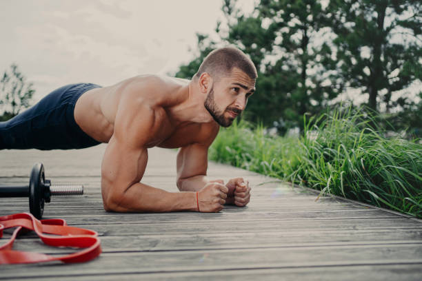 Muscular bodybuilder stands in plank pose demonstrates endurance has strong biceps poses outdoor has athletic workout, poses outdoor, keeps good physical shape. Man doing push ups, concentared forward el culturista musculoso se para en la postura de la tabla demuestra resistencia tiene posturas de bíceps fuertes al aire libre tiene entrenamiento atlético, posa al aire libre, mantiene una buena forma física. hombre haciendo flexiones, concentrado haci