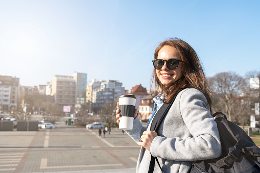 Brunette woman in sunglasses walking around the city holding backpack and reusable travel coffee cup.