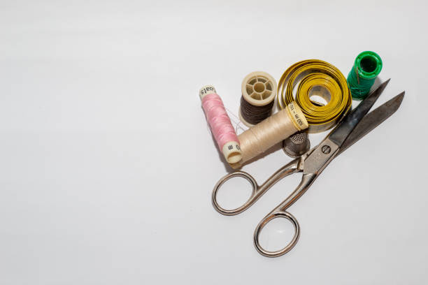 Spools of thread of different colors with a pin pinned together with a sewing scissors, a yellow tape measure, a brown button and a metal thimble on a white background.