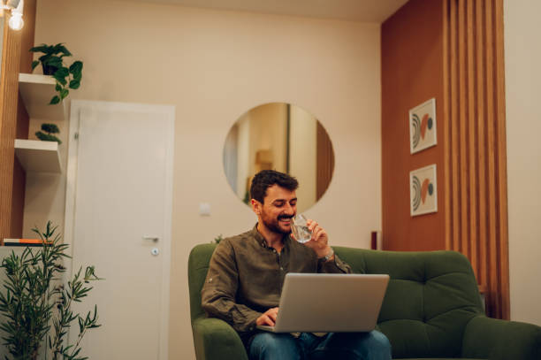 istock Caucasian bearded man using laptop and drinking water at home on a couch 1467869301