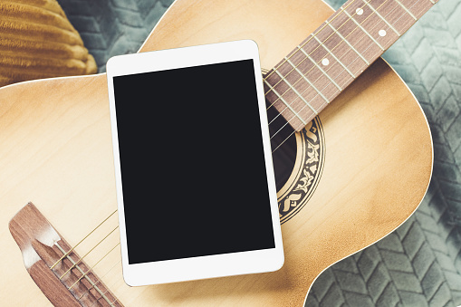 Acoustic guitar and digital tablet on couch at cozy home background. Online music lessons, learning playing or writing songs and hobby concept. Top view, flat lay, mock up