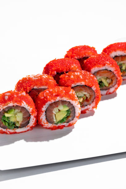 California roll with salmon, avocado and chuka seaweed, wrapped in rice with red tobiko flying fish caviar. Eight pieces stand on a white cardboard stand against a white background.