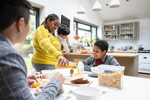 Multiracial couple in early 30s enjoying orange juice, eggs, bread, milk, and cereal with their 15 month and 10 year old sons.