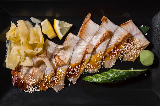 Fried eel with sauce and sesame seeds on a leaf with cucumber, wasabi, lemon and ginger on a black background