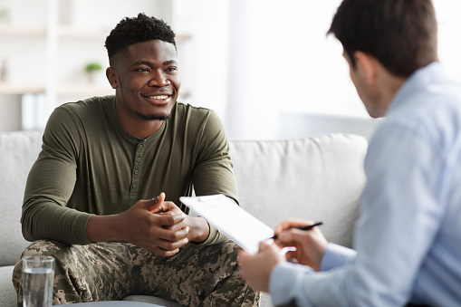 Happy young black military man in camouflage uniform sitting on couch, listening to his psychologist and smiling, enjoying results of psychotherapy. Psychological treatment for military personnel