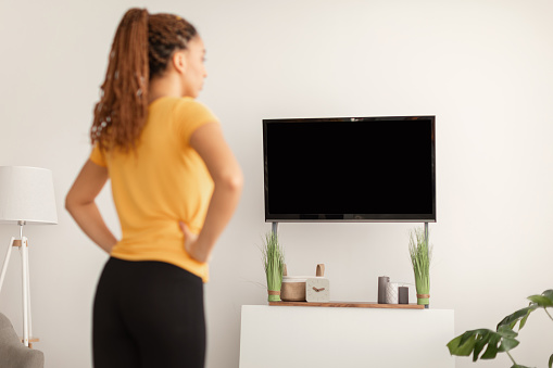 Back View Of Fitness Female Exercising Holding Hands On Hips Standing Watching Workout On Television Training At Home, Selective Focus On Empty TV Screen. Sporty Lifestyle Concept. Mockup