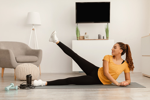 Happy African American Woman Doing Side Leg Raises Flexing Muscles Lying On Gymnastic Mat Having Workout At Home. Fitness Training And Sporty Lifestyle Concept