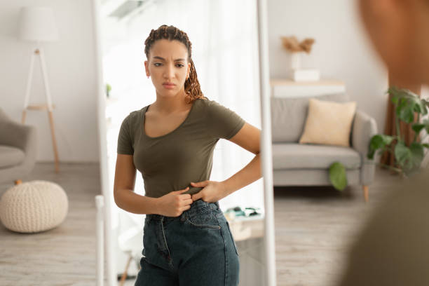 afrikanische dame berührt bauch nach gewichtszunahme in der nähe von mirror indoor - mixed age stock-fotos und bilder