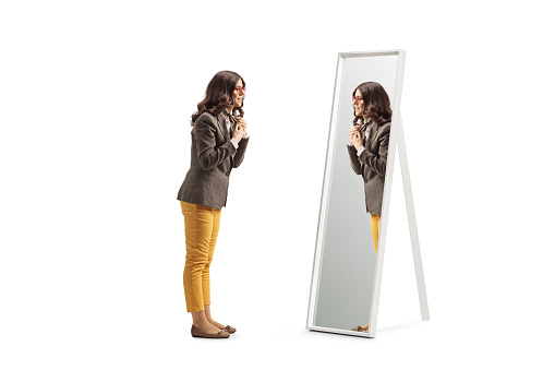 Full length profile shot of a young woman getting ready in front of a mirror and putting a scarf isolated on white background