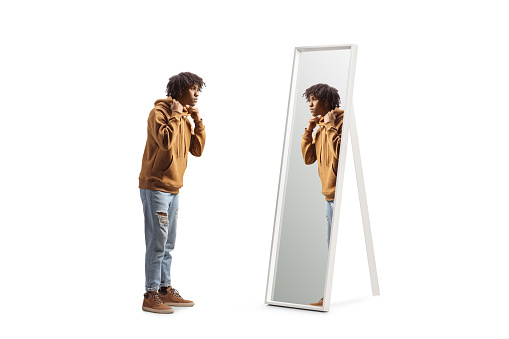 Full length profile shot of an african american young man standing in front of a mirror isolated on white background