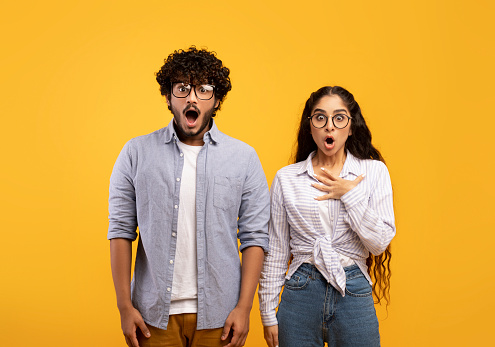 Shocked indian couple opening mouths in shock, woman putting hand on heart, standing over yellow studio background. Surprised guy and his girlfriend shouting omg, excited about huge sale or discount