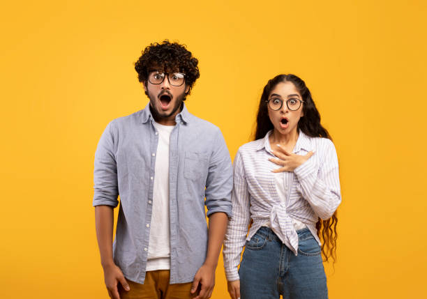 Shocked indian couple opening mouths in shock, woman putting hand on heart, standing over yellow studio background Shocked indian couple opening mouths in shock, woman putting hand on heart, standing over yellow studio background. Surprised guy and his girlfriend shouting omg, excited about huge sale or discount women screaming surprise fear stock pictures, royalty-free photos & images
