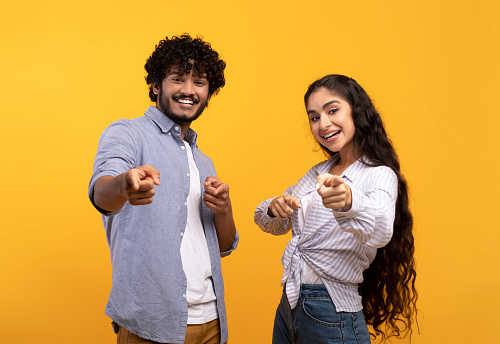 And what's about you. Excited indian couple pointing index fingers at camera, posing over yellow studio background. Positive smiling guy and lady choosing and indicating