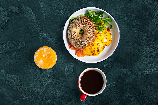 Bagel with seeds and filling of salmon and poached eggs, next to it lies a mix of salad. The food lies in a light, ceramic plate with high sides, next to it is a red cup with black Americano coffee and a transparent glass with orange juice and lion cubes.