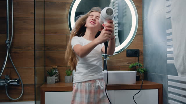 Beautiful blond little girl in bathrobe singing a song and dancing with a hair dryer. Child sings and dances in a shower after taking shower in bathroom.
