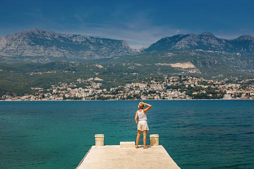 Back view of a woman tourist in straw hat standing on the edge of a pier, looking at Herceg Novi town on the other side of the bay, in Montenegro