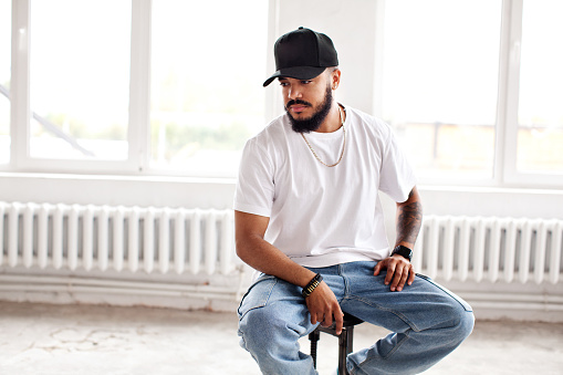 Authentic Portrait bearded man sitting in white loft background, looking away, wearing casual white T-shirt, baseball cap and jeans, lifestyle