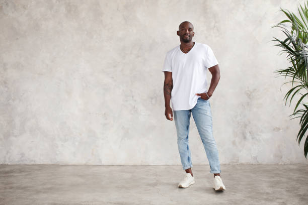 bel giovane afroamericano si erge in piedi in tutto il corpo, guarda la telecamera, contro il muro luminoso strutturato in studio. il modello maschile indossa abiti casual t-shirt bianca e jeans - male men body fashion model foto e immagini stock