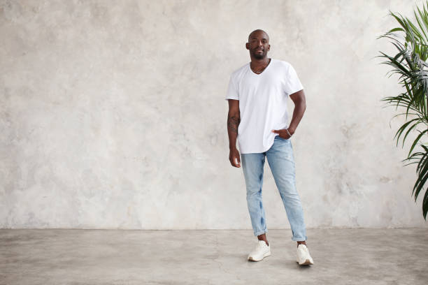 Handsome African American young man stands full body, looks at camera, against bright textured wall in studio. Male model wears casual clothes white T-shirt and jeans stock photo