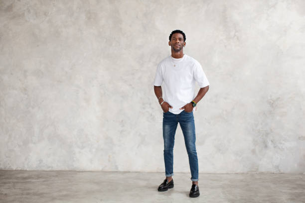 Full length African American young man stands against bright textured wall in studio. Male model keeps hands in pockets and wears casual clothes white T-shirt, jeans stock photo