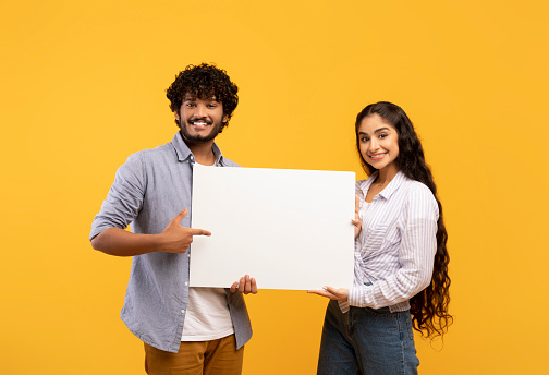 Place for your ad. Portrait of indian couple holding empty blank placard board over yellow studio background. Happy man and woman standing with white paper, pointing at it and smiling at camera