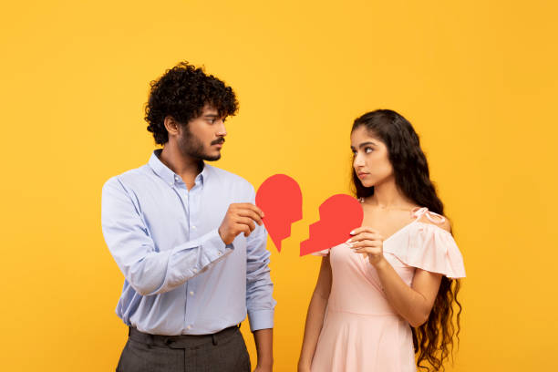 Disappointed indian man and woman holding two pieces of broken paper heart standing over yellow background Disappointed indian man and woman holding two pieces of broken paper heart standing over yellow studio background. Break up, difficulties in relationships, marriage crisis concept divorce hindu stock pictures, royalty-free photos & images