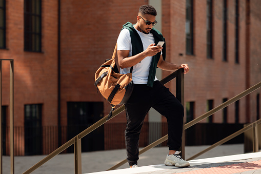 Stylish Young Black Man In Sunglasses Using Cell Phone Walking Down The Street In The Urban City, Full Body Length. Focused Trendy Guy Spending Time Outdoors. Vacation, Travel, Technology Concept