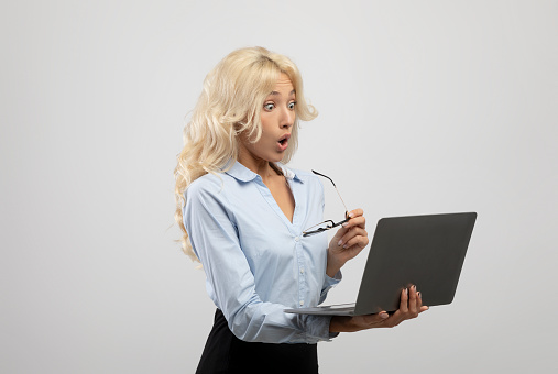 Shocked businesswoman holding laptop and looking at camera with open mouth over light grey studio background. Female office employee surprised over unexpected news, astonished at career achievements