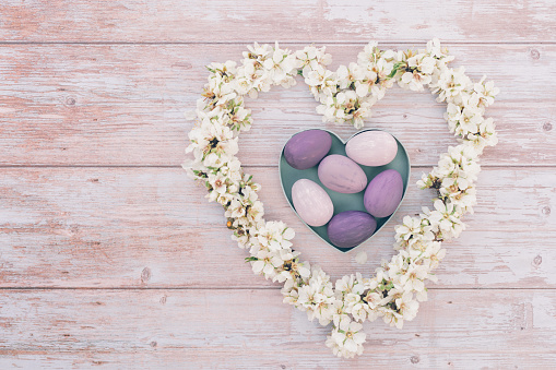 Pastel colored flat lay of pink and purple Easter eggs in heart shape box with almond blossom branches in a heart shape form on soft pink wooden Background with copy space on the left side. Color editing with added grain. Very selective and soft focus. Part of a series.