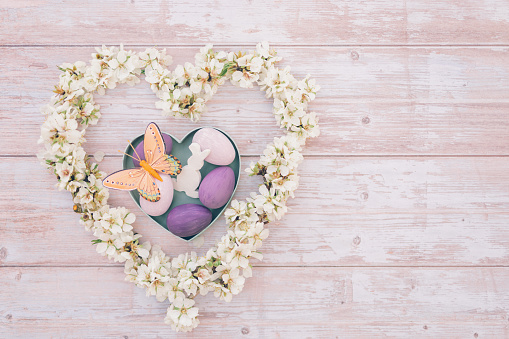 Pastel colored flat lay of pink and purple Easter eggs in heart shape box with an orange butterfly, a withe easter bunny shape and almond blossom branches in a heart shape on soft pink wooden Background and copy space. Color editing with added grain. Very selective and soft focus. Part of a series.