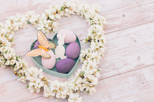 Beautiful pastel colored flat lay of pink and purple Easter eggs in heart shape box with an orange butterfly, a withe easter bunny shape and almond blossom branches in a heart shape on soft pink wooden Background and copy space. Color editing with added grain. Very selective and soft focus. Part of a series.