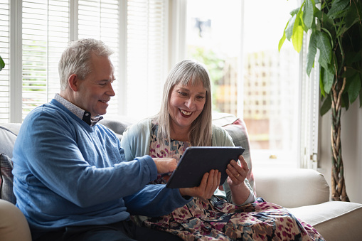 Smiling man and woman in smart casual attire sitting side by side on sofa in family home and sharing portable device.