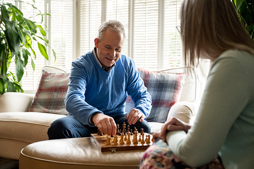 Focus on mature man in smart casual attire sitting on living room sofa across from senior woman and smiling as he moves his pawn.